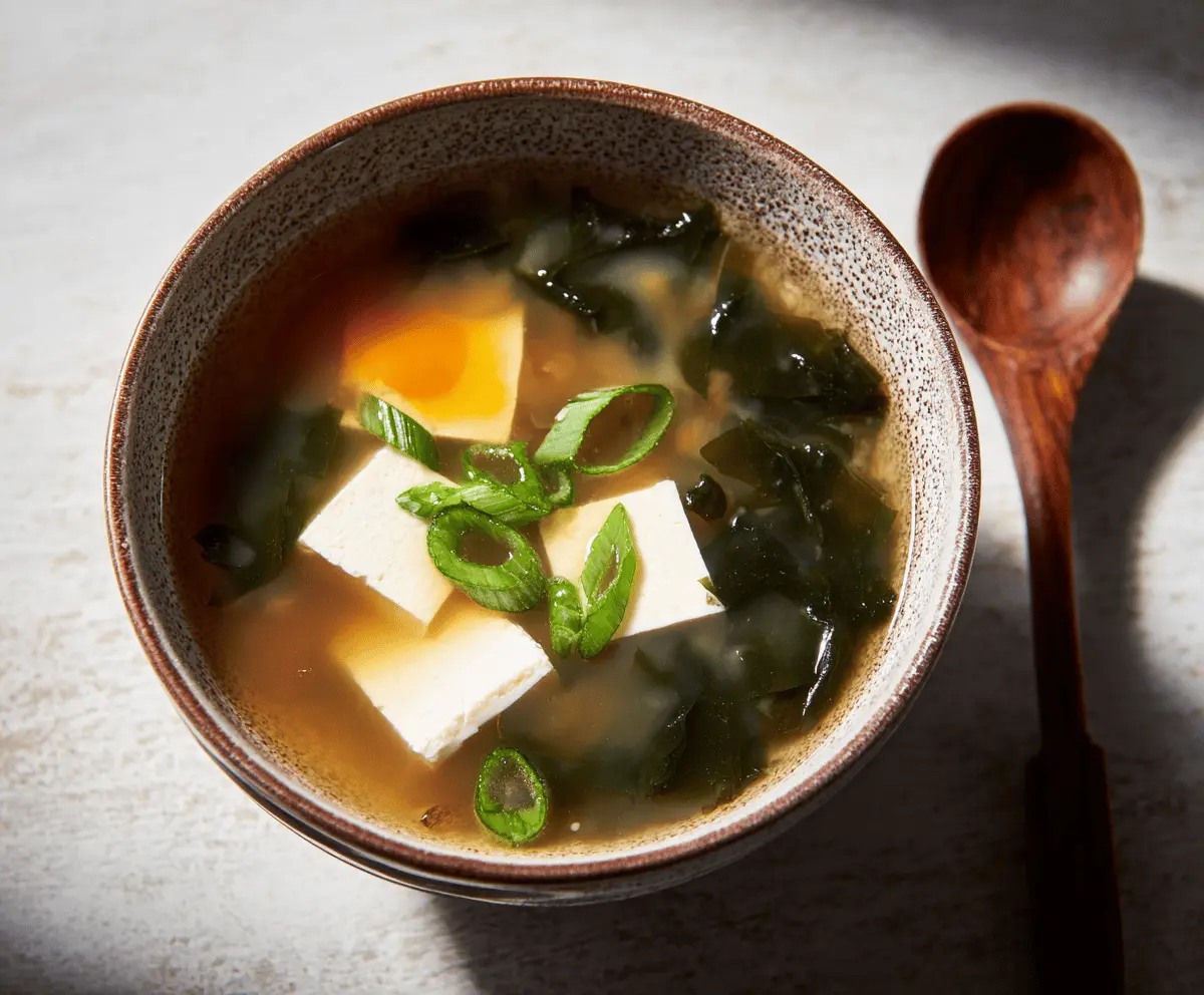 A steaming bowl of traditional miso soup garnished with sliced green onions and tofu cubes, served in a ceramic bowl on a wooden table.